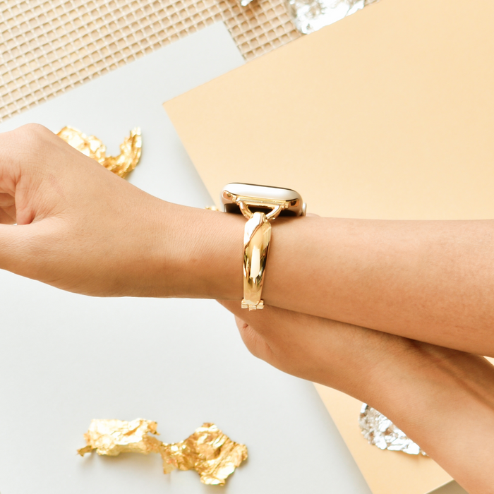 Hand adjusting a gold Coconut Lane Bangle Apple Watch Strap with gold foil and paper in the background.