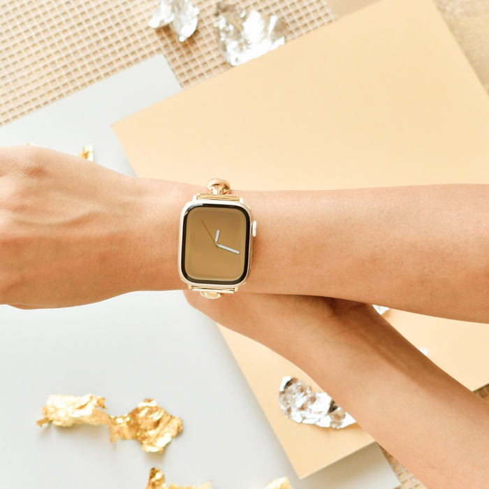 Gold Coconut Lane Bangle Apple Watch strap with beige face, worn on wrist over a beige and metallic surface.