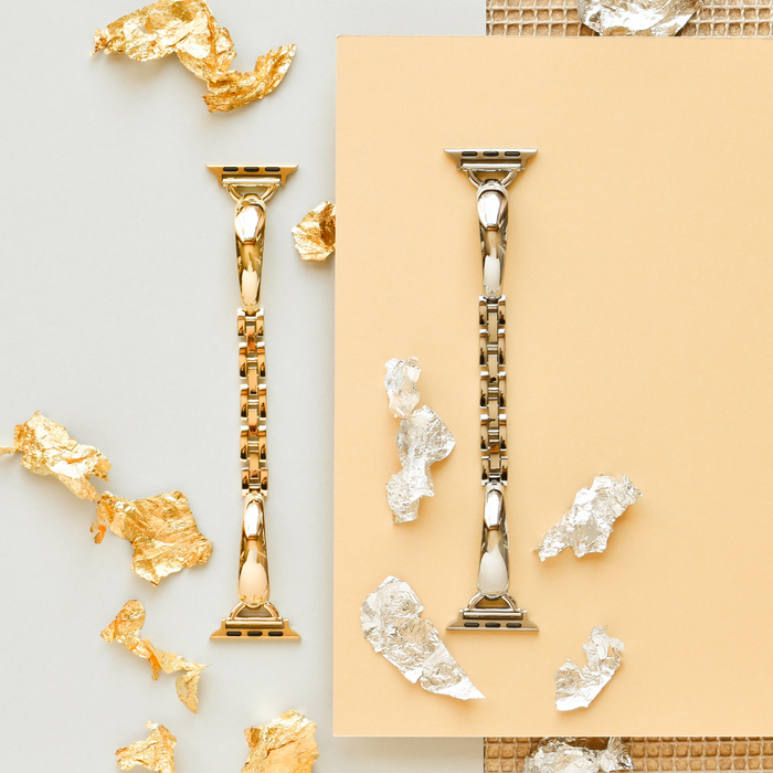 Gold and silver metal bangle-style Apple Watch straps on a beige background with metallic foil accents.