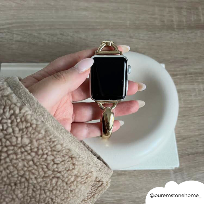 A hand holds a gold Coconut Lane bangle-style Apple Watch strap above a white dish on a wooden table.