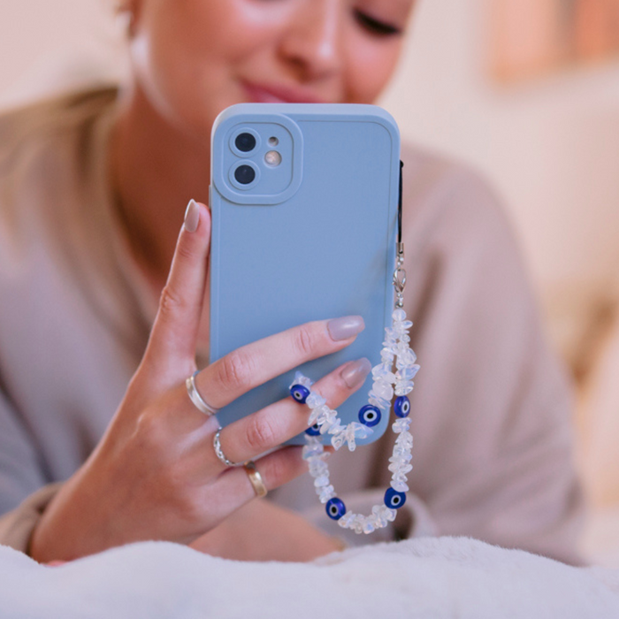 Woman indoors holding blue phone with Coconut Lane beaded phone strap featuring crystal beads and evil eye charm.