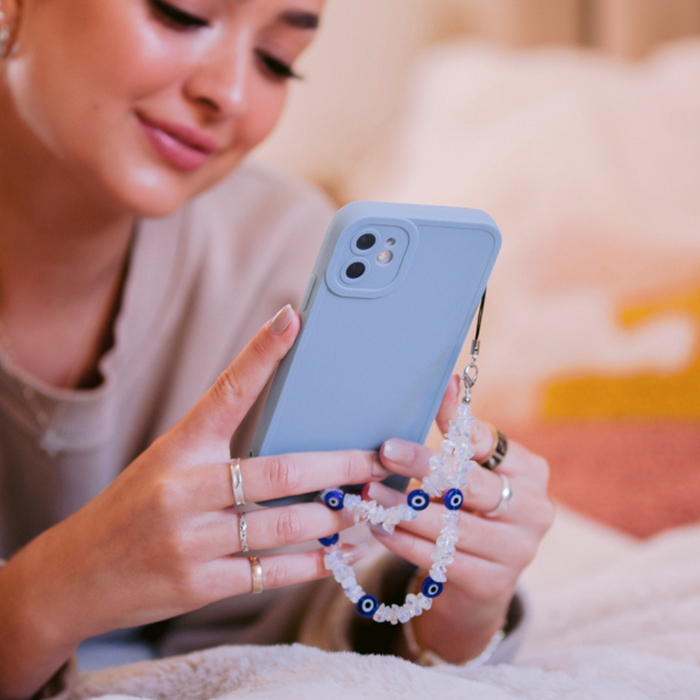 Woman on bed holding phone with blue case and Coconut Lane beaded phone strap featuring a crystal evil eye charm.