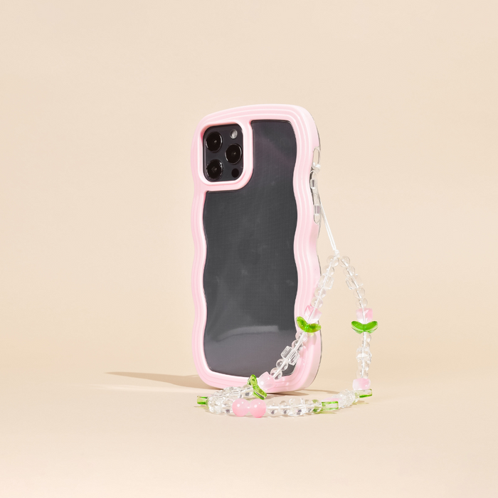 A smartphone case with a wavy pink edge, protecting a phone with triple rear cameras, is shown standing upright. Attached to the case is the Beaded Phone Strap - Cute Tulips by Coconut Lane, featuring small green leaves and pink beads that resemble cherries. This charming phone accessory stands against a plain beige background.