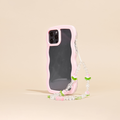 A smartphone case with a wavy pink edge, protecting a phone with triple rear cameras, is shown standing upright. Attached to the case is the Beaded Phone Strap - Cute Tulips by Coconut Lane, featuring small green leaves and pink beads that resemble cherries. This charming phone accessory stands against a plain beige background.