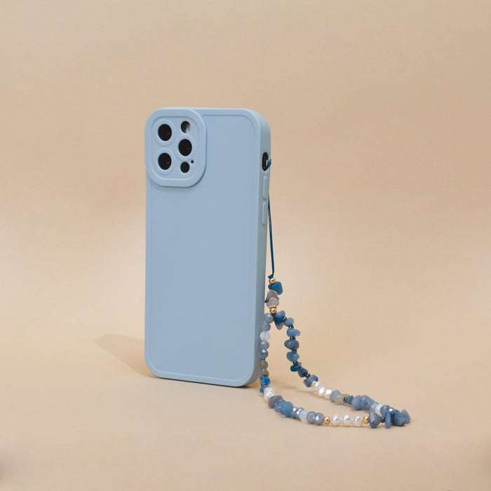 A light blue smartphone case with a camera cutout is displayed standing vertically against a beige background. Attached to the case is Coconut Lane's "Beaded Phone Strap - Ocean Eyes," made of blue beads and small pearls, adding an elegant touch to your phone accessories.