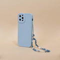 A light blue smartphone case with a camera cutout is displayed standing vertically against a beige background. Attached to the case is Coconut Lane's "Beaded Phone Strap - Ocean Eyes," made of blue beads and small pearls, adding an elegant touch to your phone accessories.