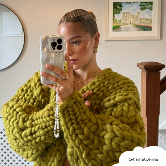 Woman in green knit sweater takes mirror selfie holding phone with pearl, gold, and pink beaded strap; art and stairs behind.