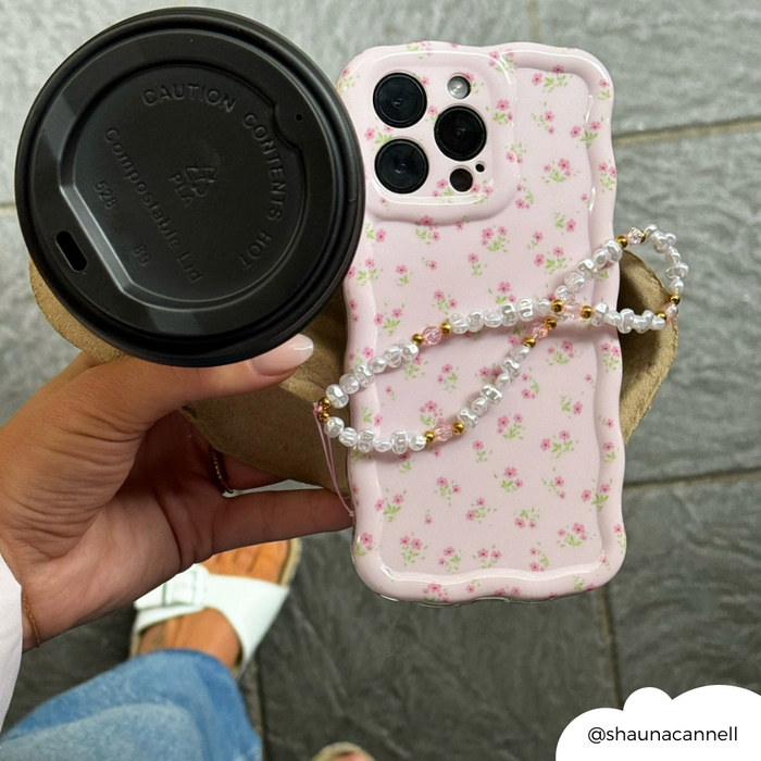 Hand holds a coffee cup and phone with Coconut Lane beaded strap in pearl, gold, and pink; jeans and shoes visible below.