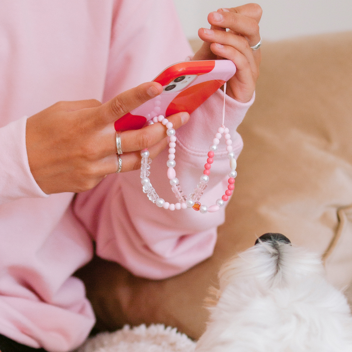 Person holding a phone with a pink beaded phone strap; small white dog looking up nearby.