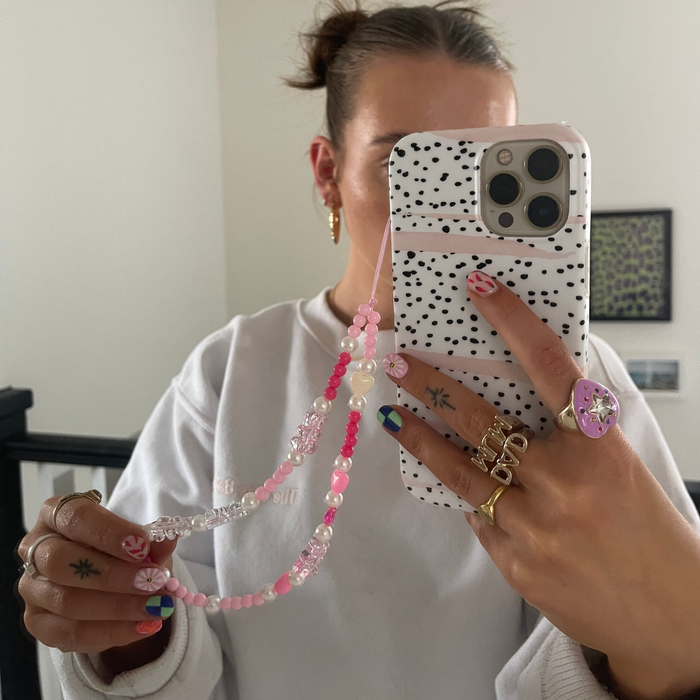 Mirror selfie showing beaded pink phone strap, colorful nails, and multiple rings on persons hand.