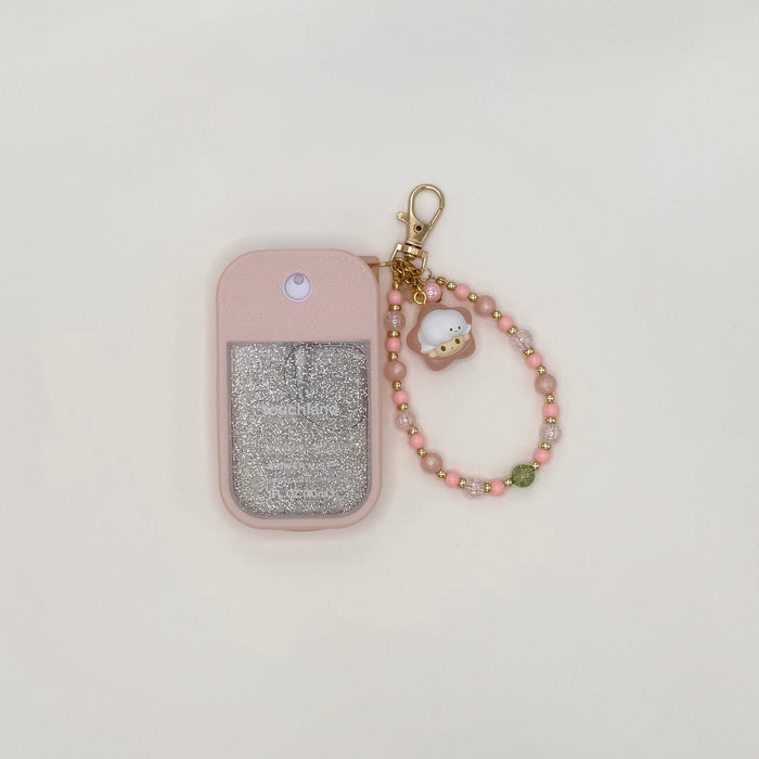 Touchland Power Mist hand sanitizer in a pink silicone case with a glitter window on a white background. Attached gold lobster clasp keychain features a Dimoo chibi charm in a yellow star hat, paired with a beaded wrist strap of pink, orange, and gold star-shaped beads with gold accent spacers. TOUCHLAND NOT INCLUDED WITH THIS PRODUCT!