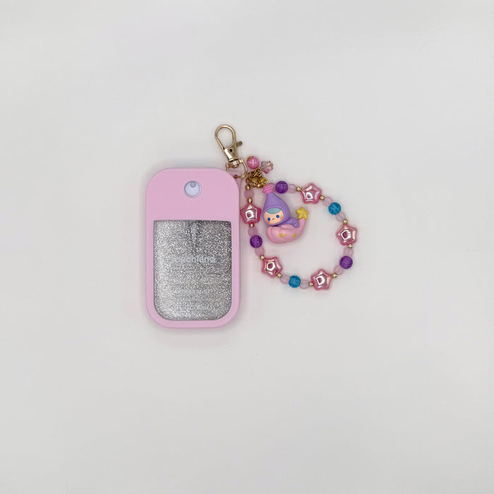Touchland Power Mist hand sanitizer in a pink silicone case with a glitter window, styled on a lavender bow bag. Attached charm features a Pucky chibi figure in a purple flower hood with blue accents, dangling from a beaded strand of purple glitter, lavender, and gold beads with a pink ball accent. TOUCHLAND NOT INCLUDED WITH THIS PRODUCT!
