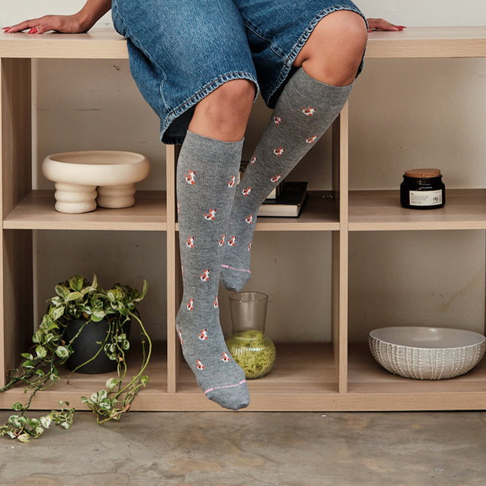 Knee-high gray compression socks with cat patterns, worn by a person sitting on a wooden shelf.