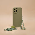 A Crystal Phone Strap - Green by Coconut Lane is displayed on a beige background, showcasing its beaded charms attached to a green phone case. Small green stones and a polished Green Aventurine gemstone are scattered beside it, creating a minimalist and earthy aesthetic.