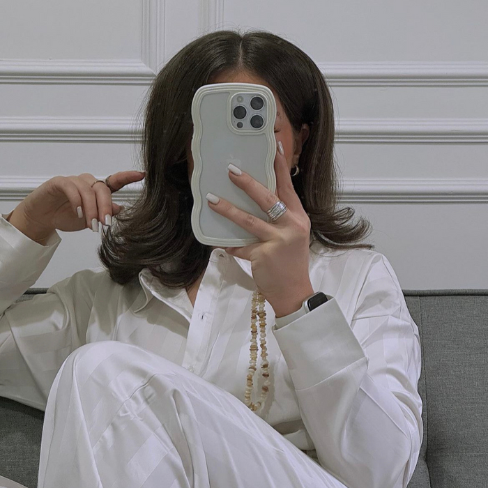 Woman in white pajamas sits on a couch, holding phone with Coconut Lane natural nude crystal phone strap.
