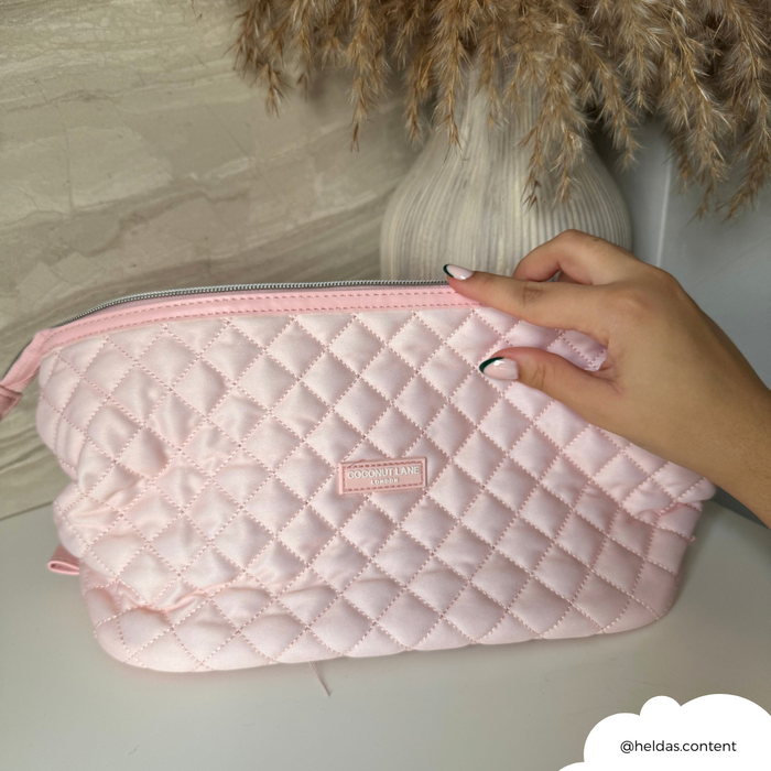 Hand holding a pink quilted Coconut Lane expandable makeup bag on a white surface, vase in background.