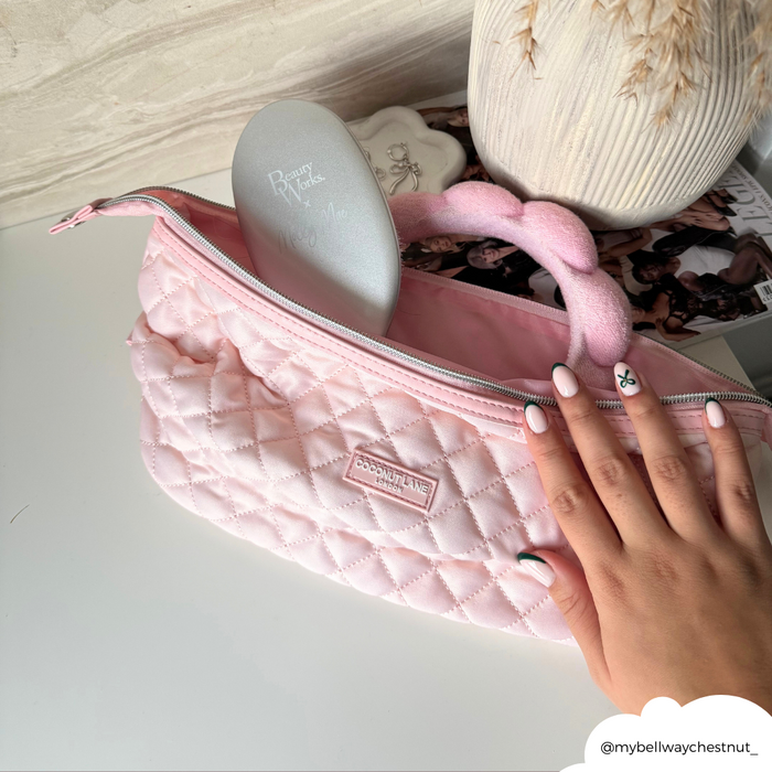 Hand with nail art on a quilted pink Coconut Lane makeup bag, showing a hairbrush and scrunchie inside.