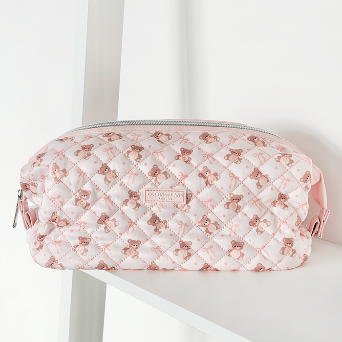 Pink quilted makeup bag with teddy bear pattern and zipper, displayed on a white shelf.