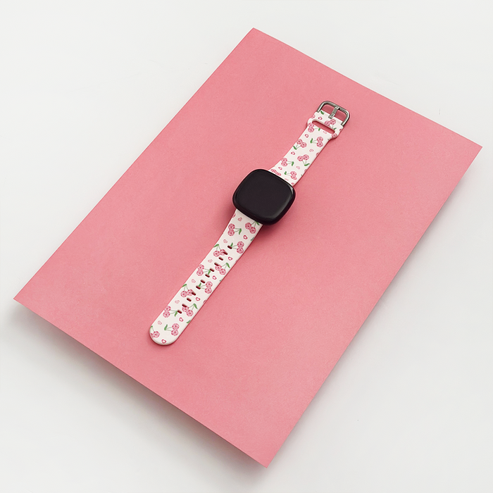 Coconut Lane Disco Cherries Fitbit strap with cherry pattern on white, displayed on a pink surface.
