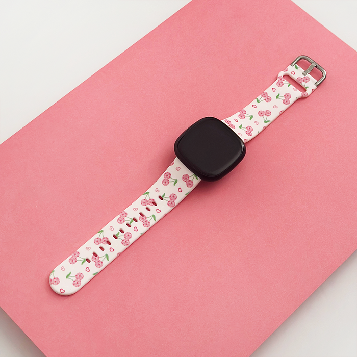 Fitbit strap by Coconut Lane with red cherry and heart print, displayed on a pink background.