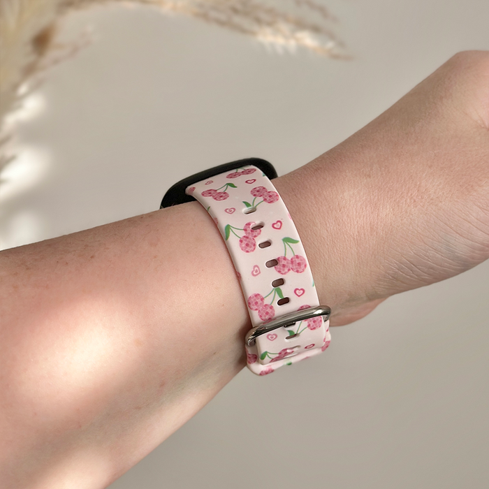 Pink silicone Fitbit strap with cherry and heart pattern, worn on wrist.