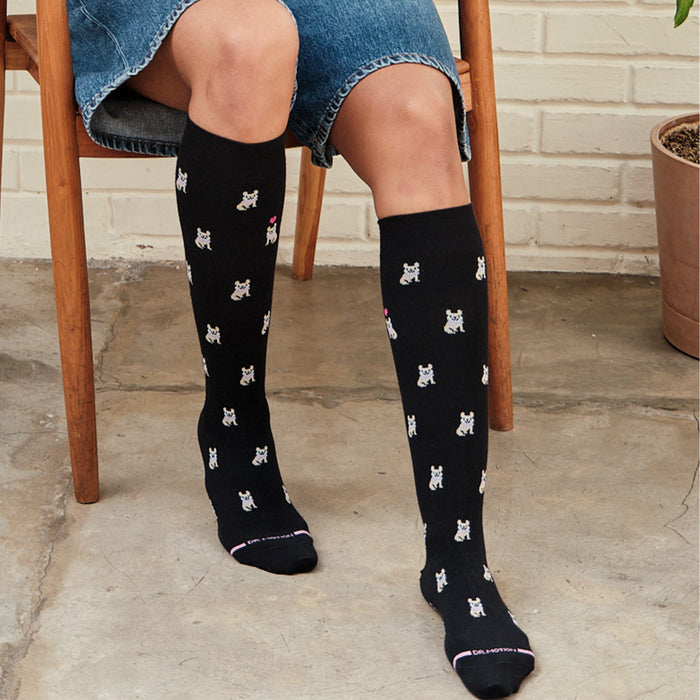 Knee-high black compression socks featuring a French Bulldog pattern, worn by a person seated on a chair.