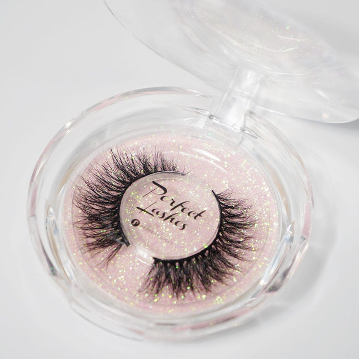 A clear, round plastic case displays dramatic, thick false eyelashes labeled Perfect Lashes on a sparkling, glittery pink background.