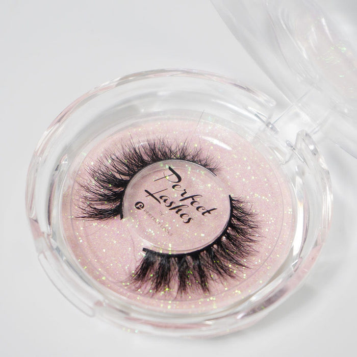 A pair of thick, dramatic false eyelashes in a round clear case, sparkly pink background, Perfect Lashes label on the lid.