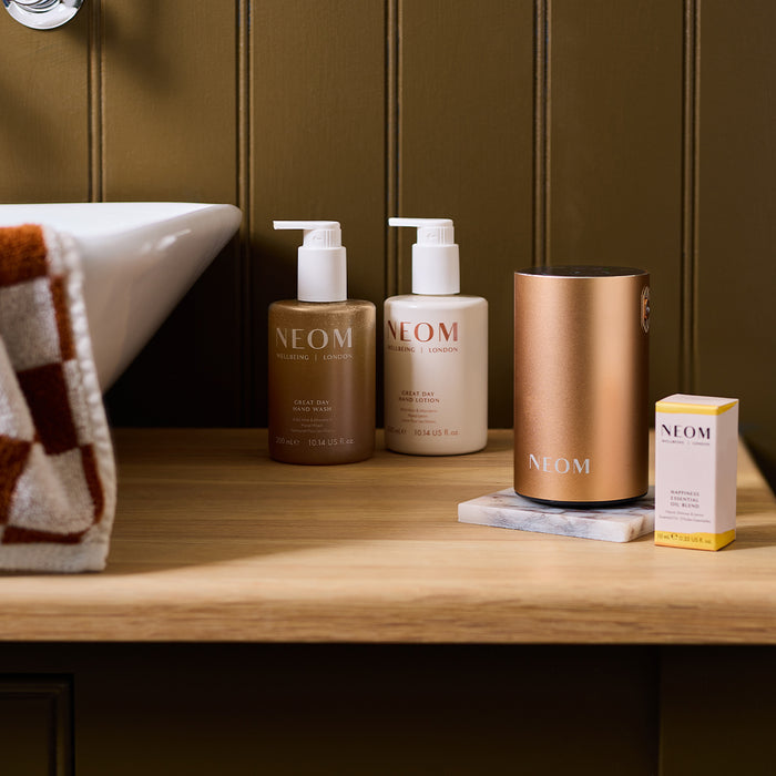 NEOM pod mini+ in rose gold, essential oil blend, hand wash and hand lotion in a a bathroom beside a sink