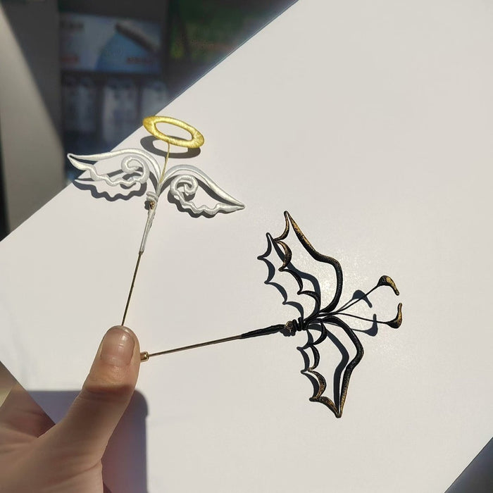 Handheld view of a pair of handmade Chanhua thread-wrapped brooches featuring a white angel wing with a gold halo and a black demon wing with gold tips.