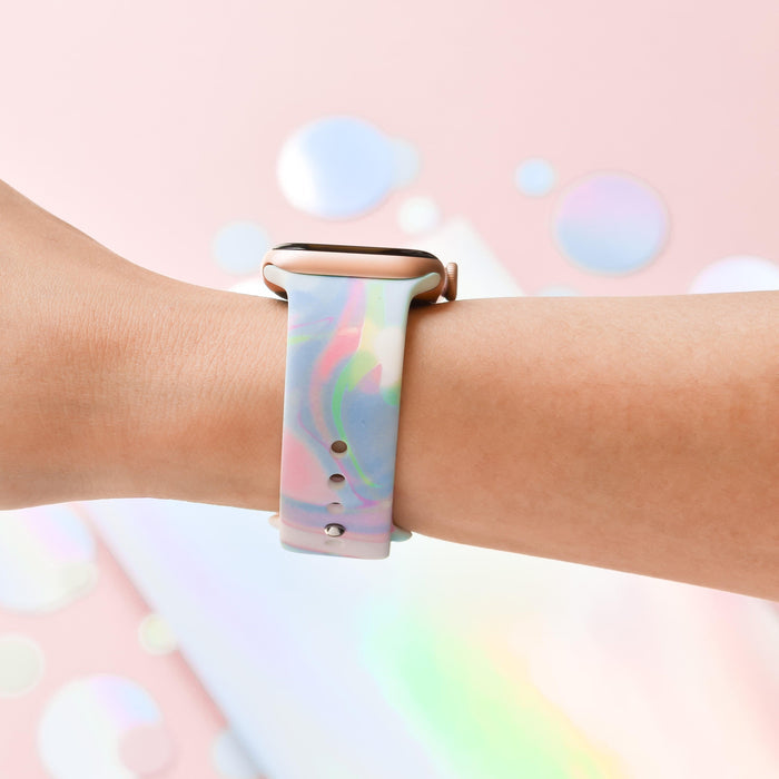 A close-up image of a person's arm wearing an Apple Watch paired with the Coconut Lane Holo Bubble Apple Watch Strap, showcasing its multi-colored, pastel gradient. The background is a soft pink with blurred circular reflections that create a dreamy, abstract atmosphere, perfectly accentuating the strap's holographic iridescent design.