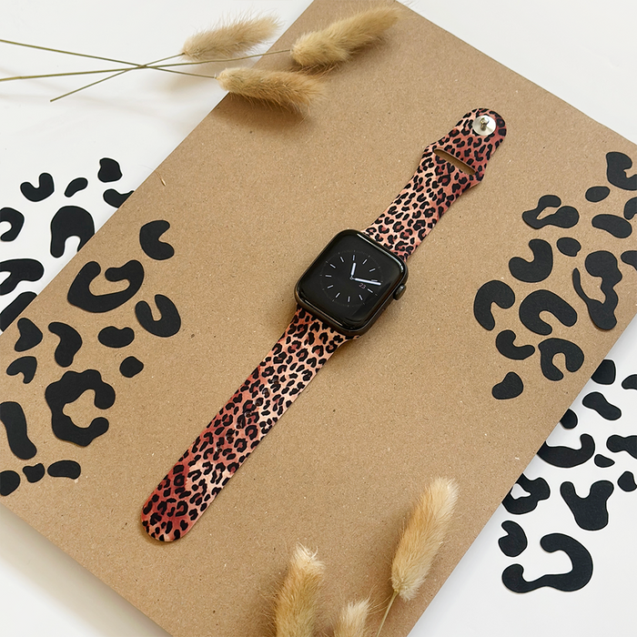 Leopard print Apple Watch strap with black animal spots, displayed on a brown board with dried grass decoration.