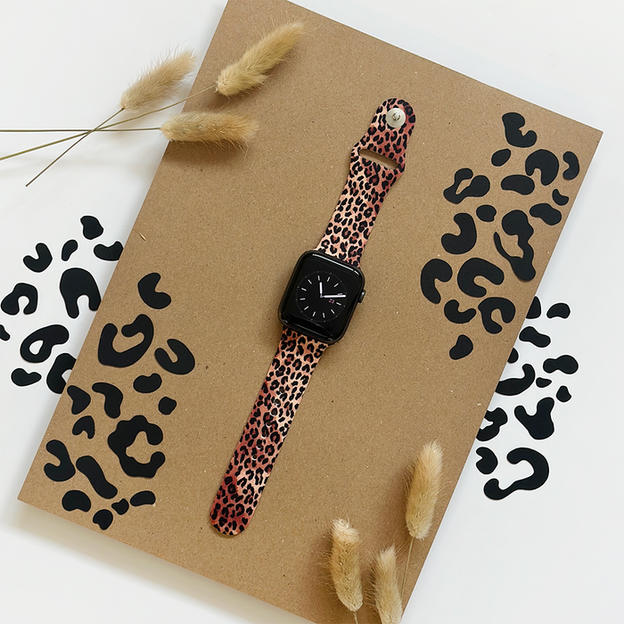 Leopard print Apple Watch strap with black spots on brown, displayed on a wooden board with dried grass accents.