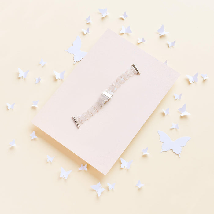 The Luxe Holo Butterfly Apple Watch Strap by Coconut Lane is showcased on a light beige rectangular board, surrounded by scattered holographic butterflies of various sizes. The white beaded strap creates a delicate and airy composition against the soft, pale yellow background.