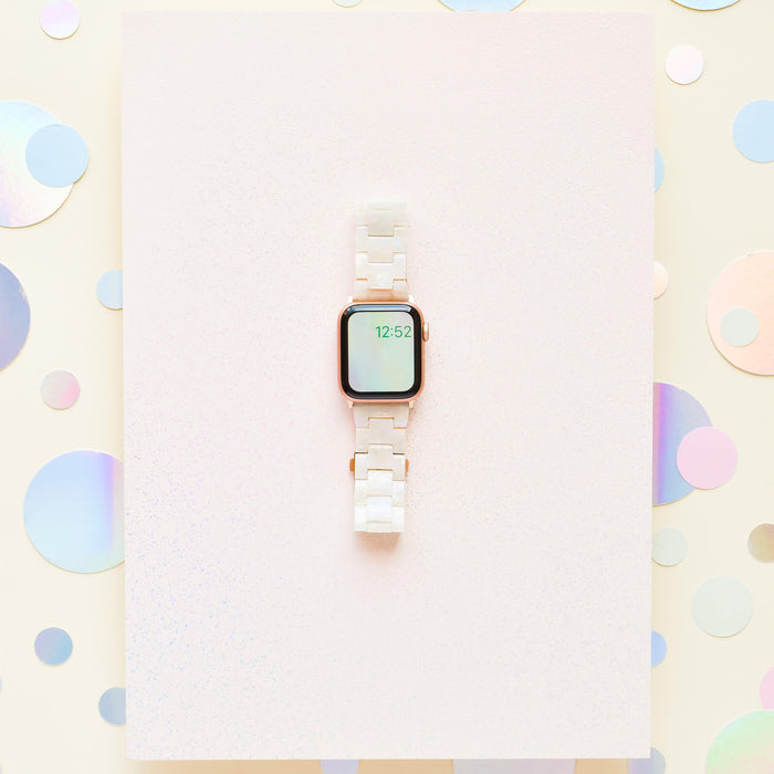A smartwatch featuring the Luxe Iridescent Apple Watch Strap from Coconut Lane, with a white ceramic band, is displayed against a pastel background with holographic dots. The watch screen shows the time as 12:52. The minimalist and modern design perfectly complements the stylish and chic setup, further enhanced by hints of the iridescent watch strap.
