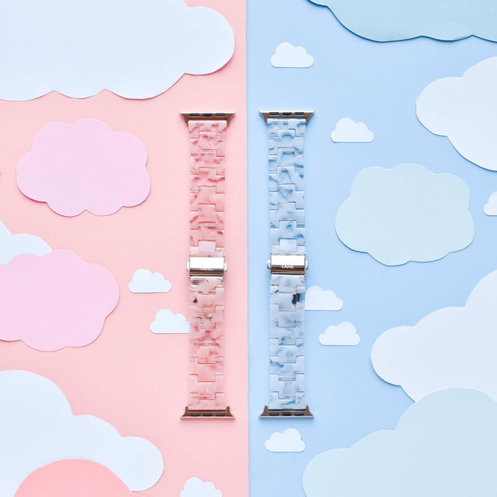 An image features two Apple Watch straps from Coconut Lane, including the Luxe Pink Clouds Apple Watch Strap and a blue counterpart, placed vertically side by side on a background divided into corresponding pink and blue sections. Each section is adorned with matching paper clouds, creating a girly and cute atmosphere. These durable resin bands add both style and function to your device.