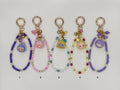 Five numbered kawaii wrist strap keychains on a white background, each with a gold spring ring clasp and a macaron-shaped dome charm containing a chibi character face. Variants include a purple strap with blue bunny charm (1), pink and mauve strap with pink piggy charm (2), white and blue strap with yellow Alice-style charm (3), yellow-green and red strap with pink chibi charm (4), and an all-purple strap with blue character charm (5).