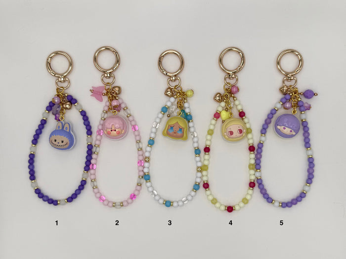Five numbered kawaii wrist strap keychains on a white background, each with a gold spring ring clasp and a macaron-shaped dome charm containing a chibi character face. Variants include a purple strap with blue bunny charm (1), pink and mauve strap with pink piggy charm (2), white and blue strap with yellow Alice-style charm (3), yellow-green and red strap with pink chibi charm (4), and an all-purple strap with blue character charm (5).