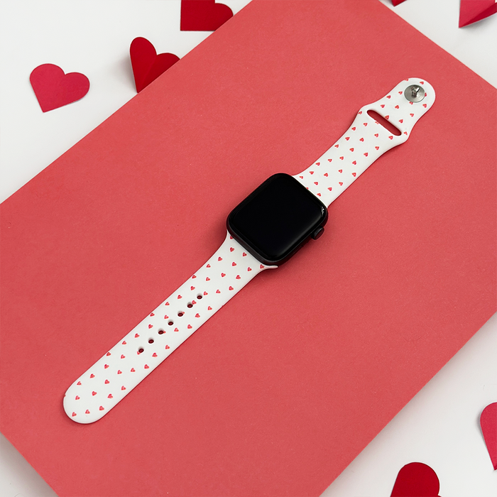 Pink Apple Watch strap with small heart pattern, displayed on a pink surface with paper heart decorations nearby.