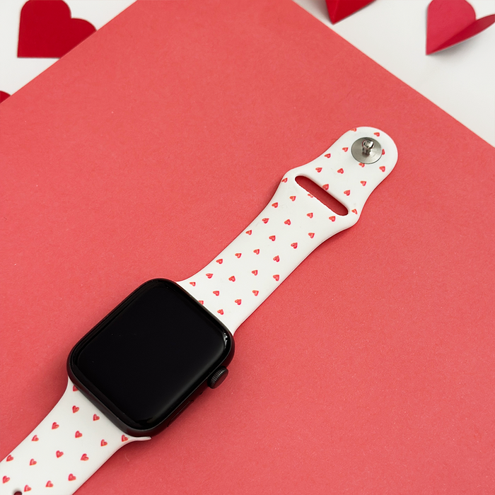 White Apple Watch strap with small heart pattern, displayed on a pink background with paper hearts.