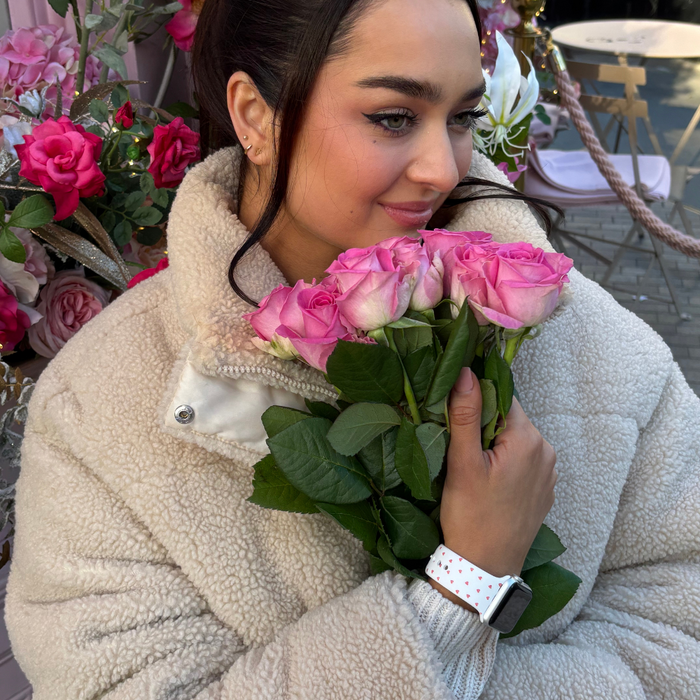 Woman in a beige coat holds pink roses, wearing a white Apple Watch strap with small red heart pattern.