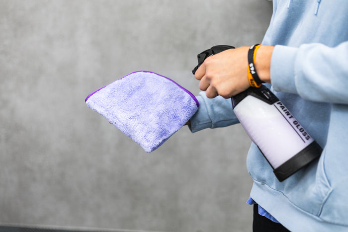Person holding a white iK spray bottle with a purple minx royale microfiber cloth against a gray background