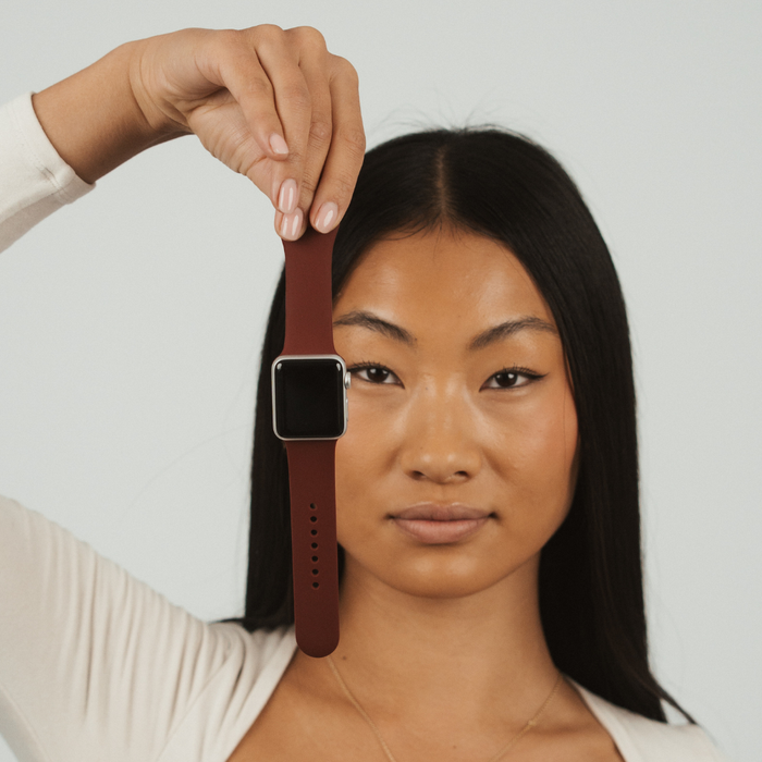 Woman holding a burgundy NAKD Apple Watch strap by Coconut Lane in front of her face, plain background.