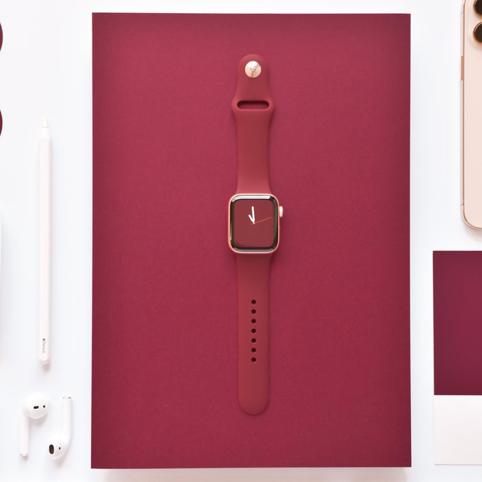 Burgundy Apple Watch strap by Coconut Lane with white and gold accessories on a matching burgundy background.