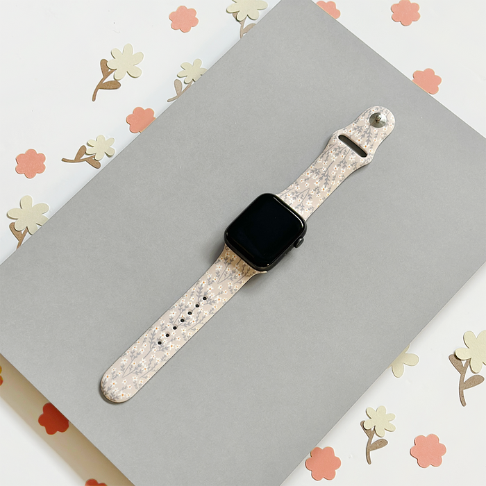 Coconut Lane Apple Watch strap in nude with floral pattern, shown on a gray surface with flower accents nearby.