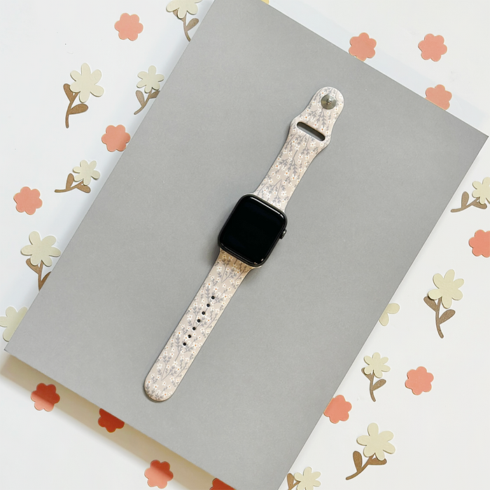 Apple Watch strap in nude with white floral pattern, shown on a textured background.