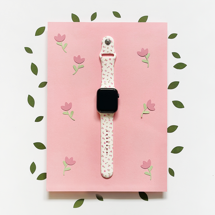 Pink watercolour floral Apple Watch strap on a pink sheet, surrounded by green paper leaves.