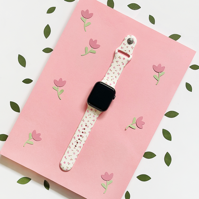Pink floral Apple Watch strap with watercolor flower pattern, shown on a pink background with green leaves.
