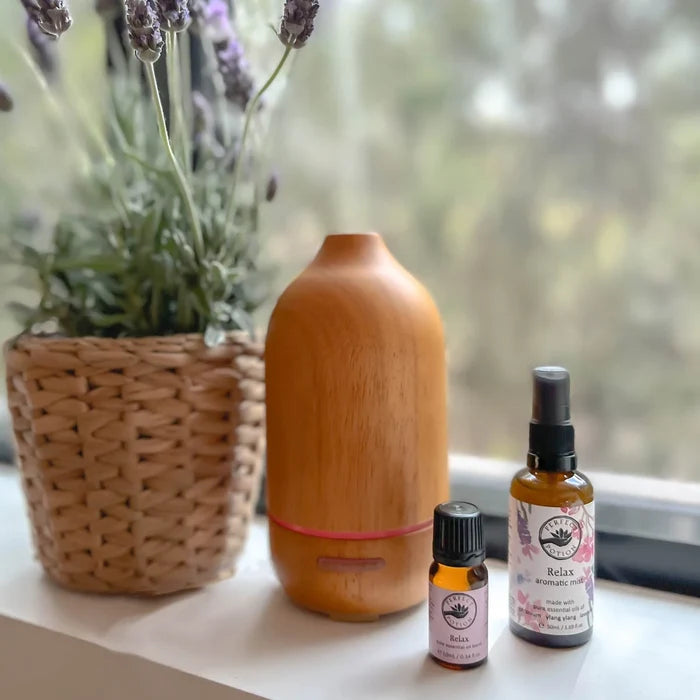 A wooden diffuser bottle stands on a windowsill next to a woven basket planter with lavender and two small bottles of Relax aromatherapy products.