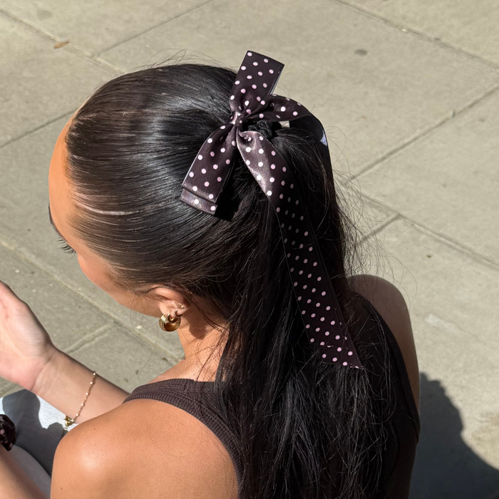 Woman with dark hair wears a brown polka dot ribbon bow clip while sitting on a sunlit sidewalk.